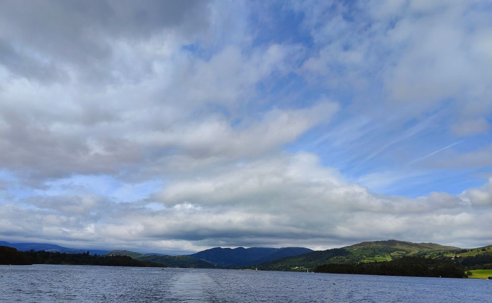A shot from Windermere. The boat's wake is visible in the water.  The sky takes up most of the picture life white clouds dissolve into wisps. The land is a bunch of nested hills.