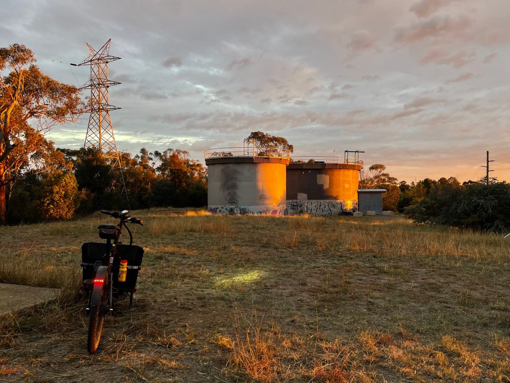 A photo, taken in the evening, with a cargo bike, a power line and 2 large water tanks. The sky is overcast which contrasts against the golden glow of the evening light. 