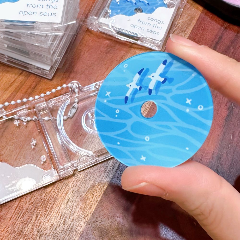 A tiny CD charm design that shows the ocean and seagulls! The CD case is open and I am holding a tiny ocean designed CD in my fingers. 