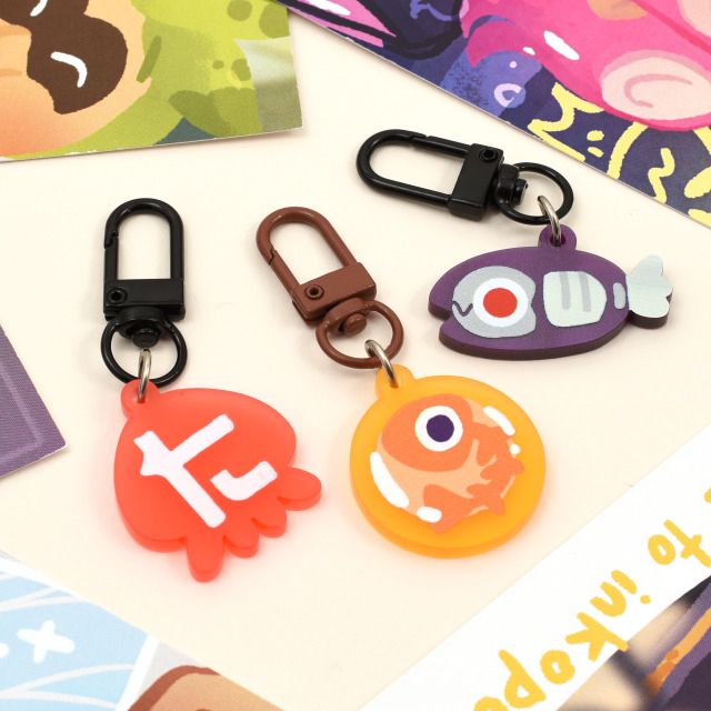 Photo shows 3 Splatoon charms made by me! One is a salmon egg design on soft frosted yellow acrylic, one a red squid with the logo for the Toni Kensa splatoon brand on it, and the second is a small Jelleton fish enemy from Splatoon Side Order. They are placed on a surface together with other Splatoon items!