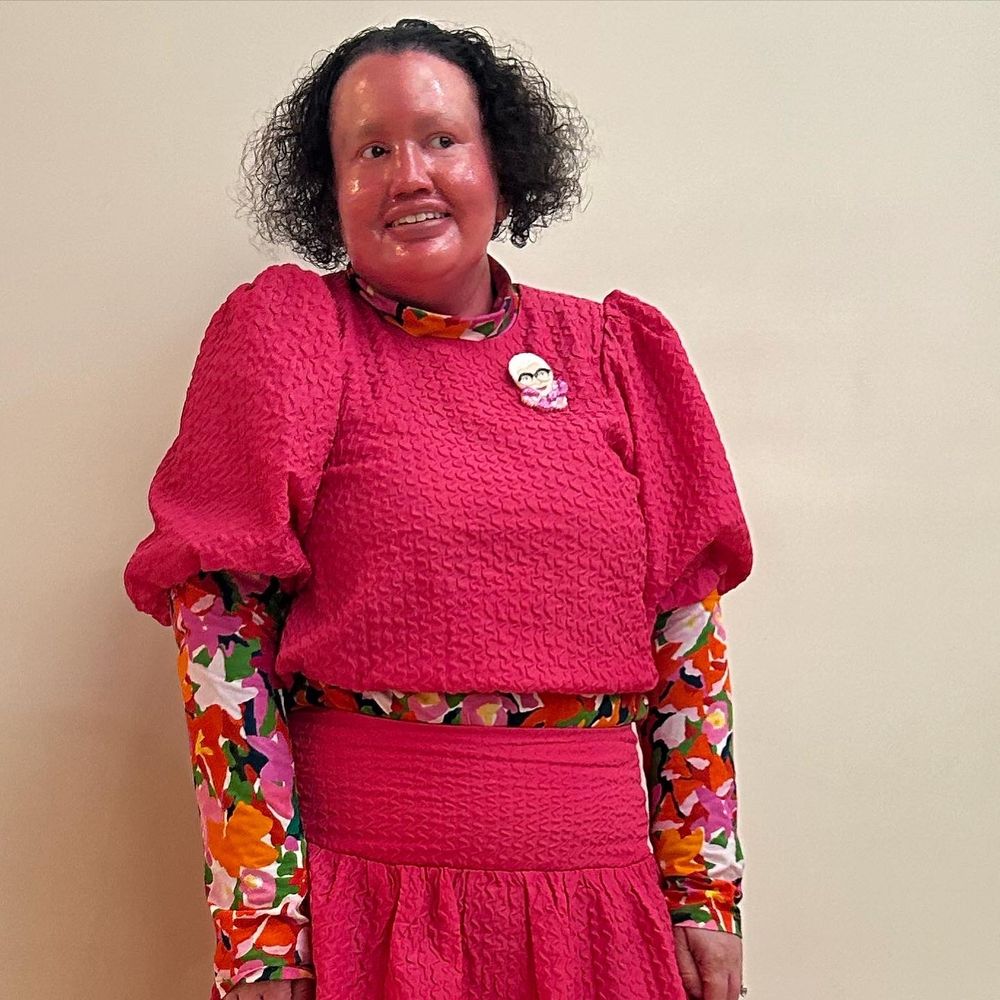 Carly, a woman with a red face, short dark curly hair, wearing a puffed sleeve top and long ruffle skirt, both are hot pink. The top is worn over a colourful floral skivvy. She also has chunky black boots on, and a brooch that is Iris Apfel's face. She's standing in the Melbourne Recital Centre foyer. The first photo is full length, the second is up close.