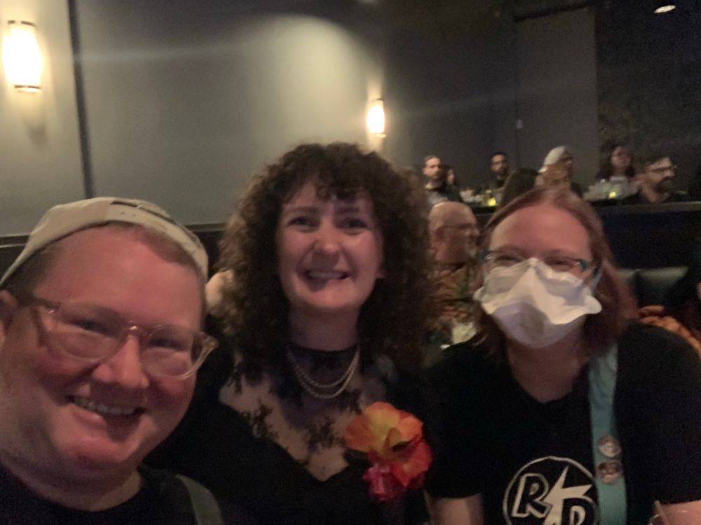 Three people inside a darkish room smile at the camera. One is wearing a baseball hat backwards one has curly hair and the other is wearing a mask and glasses. They are at a comedy club about to see RHYS DARBY!!