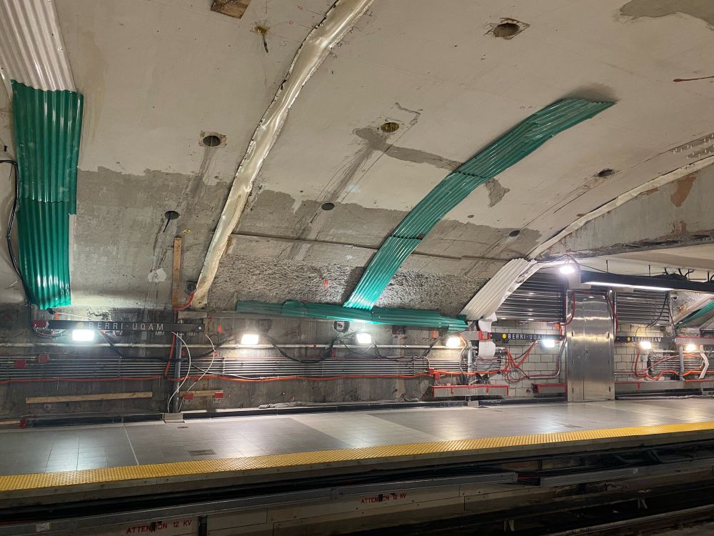 Berri UQAM station, yellow line. Exposed concrete, red-orange tubing, kelly green corrugated metal patched in a line up the wall.