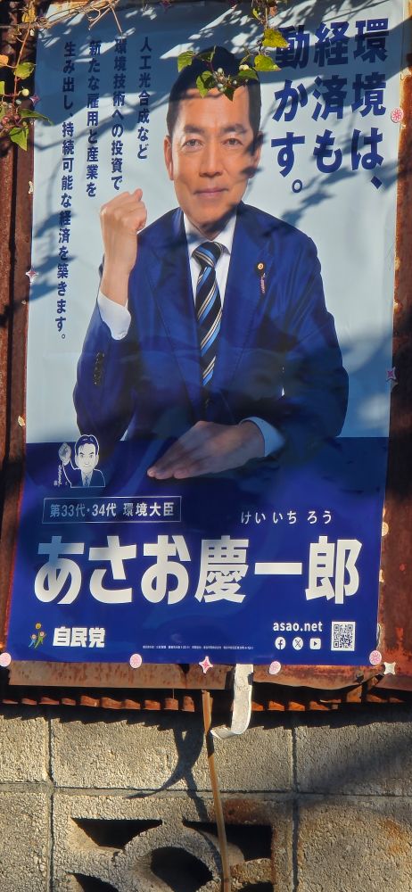 A poster for Keichi Chirro. He is sitting at a table with his shit eating grin. He has just right fist up. He looks like he is explaining why he had to poison the water supply for the local elementary school to lower property prices so he could build a factory cheaply. He has the same life in his eyes as a gutted fish.