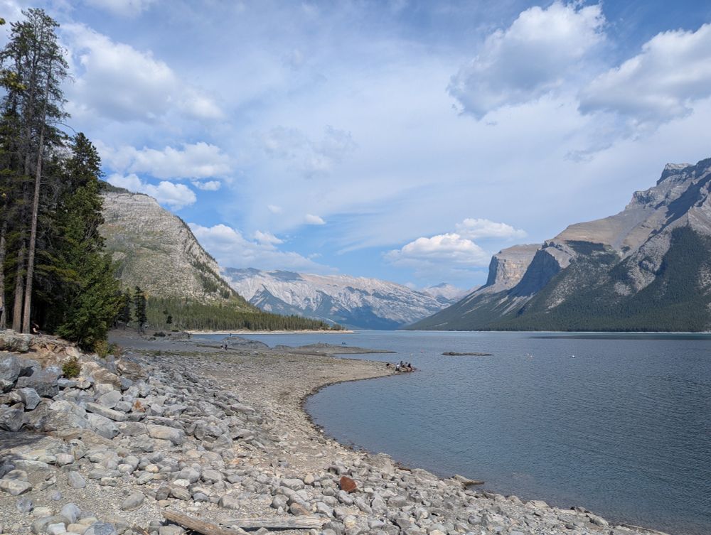 View of a rocky lake shore with the lake surrounded by mountainous terrain and pine trees.