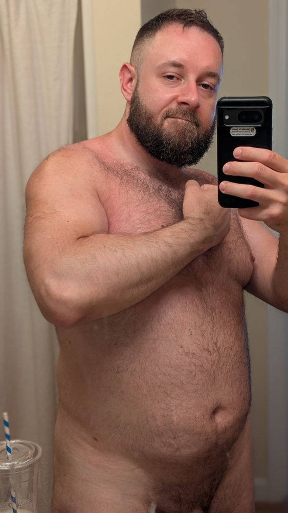 Beardy bearish man, naked, looking into the camera, shot is framed from the dick root up, he's flexing kinda silly and looks meaty 