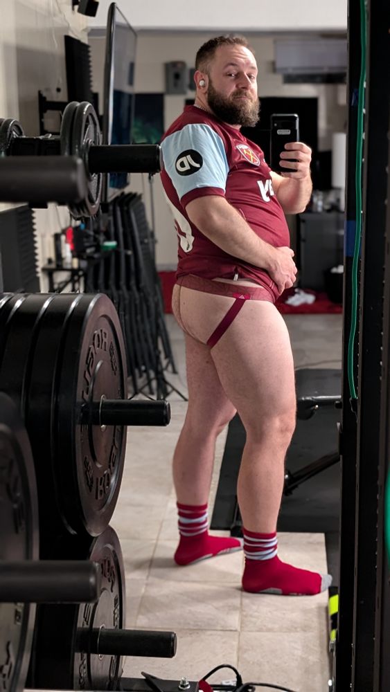 Beardy bearish man wearing a UK soccer jersey, with matching maroon jockstrap and socks