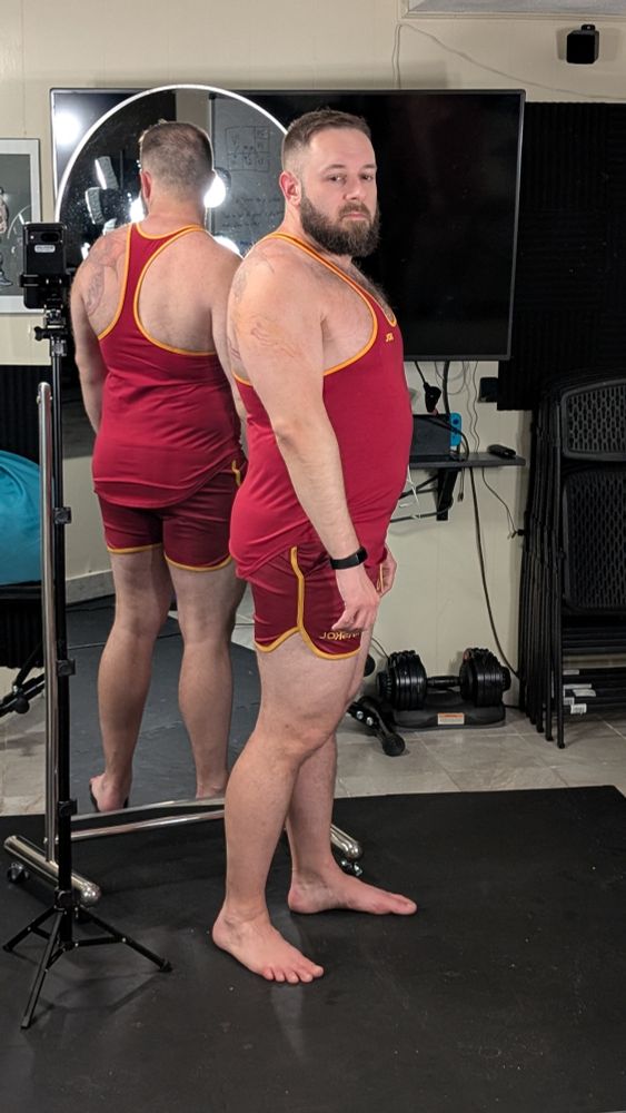 A beardy bearish man, wearing a black fitbit, standing in front of a mirror, with a mirror behind him, taking photos in a Raskol tank top and matching shorts