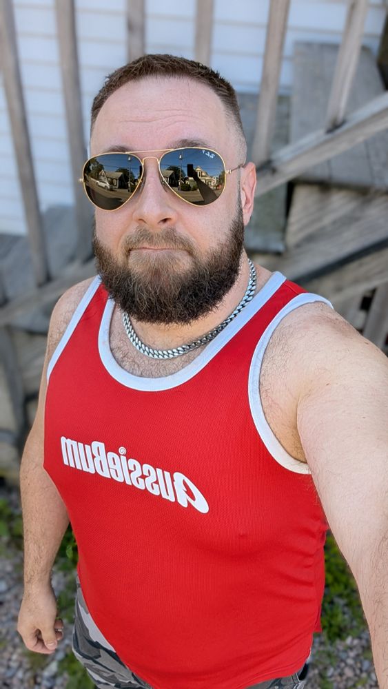 Beardy bearish man in a red Aussiebum tank top and aviator sunglasses, with a silver chain