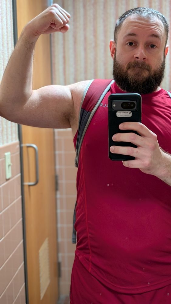 Beardy bearish man standing in front of a mirror taking a photo, while flexing his left arm
