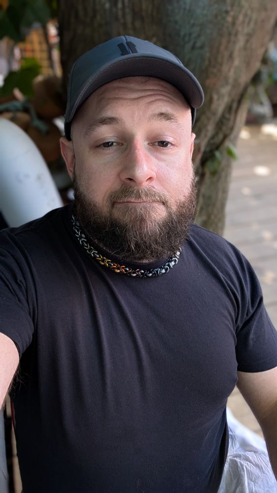 Beardy bearish man wearing a black t shirt, gray light camo hat, and a chain showing bear pride colors 
