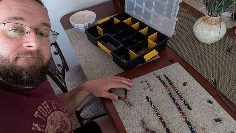 A beardy bearish man with two sets of pliers and some chain links, showing a partially completed bear pride chain, a finished rainbow pride chain, and a bear pride bracelet