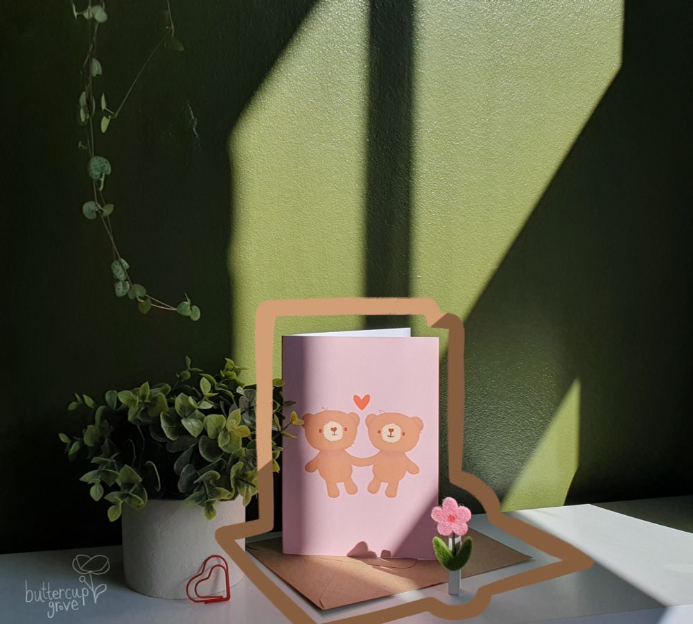photograph of a green wall and a white shelf in a sunny room with a plant and a greetings card sat on the shelf. the greetings card is pink with two smiling brown bear soft toys holding hands with a red heart above them. in the bottom left is a semi transparent watermark of the buttercup grove logo, the handwritten name and a simple buttercup drawing to the right of the words. drawn on top of the photo around the shape of the card and envelope is an orange and brown coffee coloured line. in the bottom left corner is a watermark of buttercup grove and a simple buttercup logo.