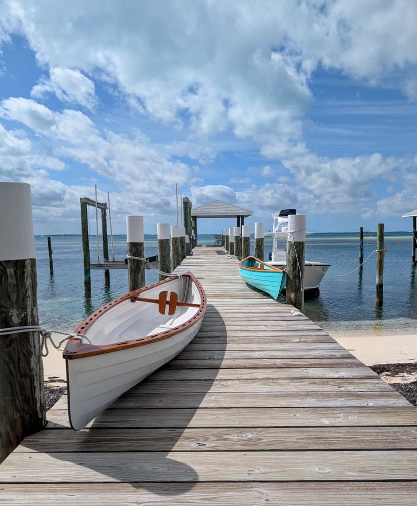 A sunlit wooden dock stretches into a turquoise sea under a sky of fluffy white clouds. Two small, elegant boats, one cream and one teal, rest peacefully on the dock.