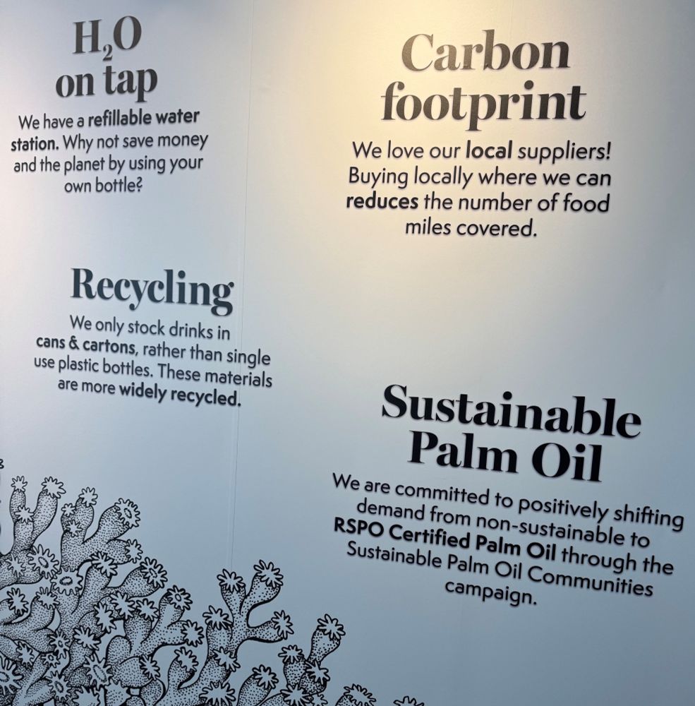 A wall display promoting environmental sustainability, highlighting themes such as water refilling, carbon footprint, recycling, and sustainable palm oil use. The display includes brief descriptions of initiatives like using refillable water bottles, supporting local suppliers to reduce travel distances for goods, promoting recycling through eliminating the use of single-use plastics, and a commitment to sustainable palm oil. The background is textured and the text varies in size to emphasize different points.