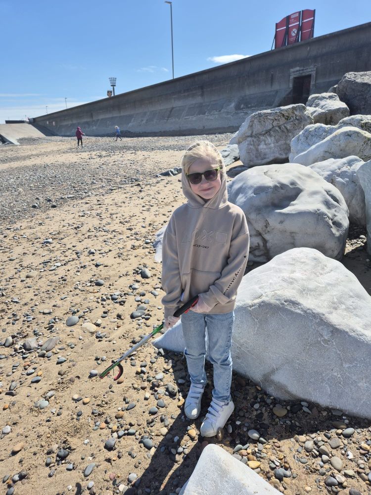 A child smiling while standing on a pebbly beach with large white rocks, holding a litter picker near a seawall under a clear blue sky.