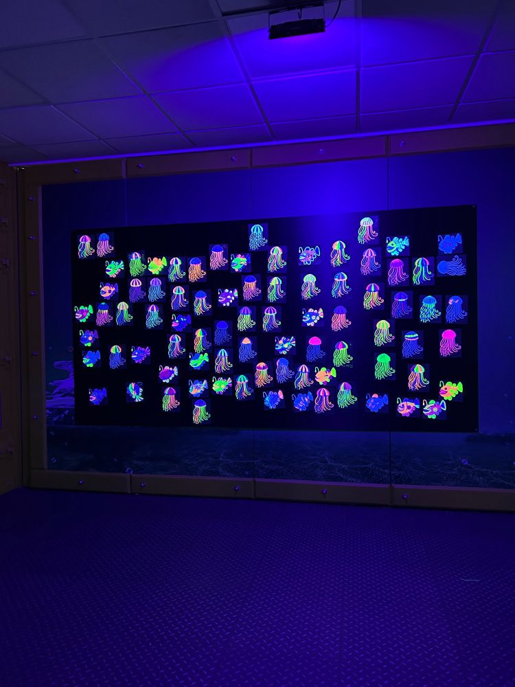 A big black wall filled with a range of colourful illuminated UV jellyfish and anglerfish, that have been coloured in and stuck to the glow wall by visitors. 