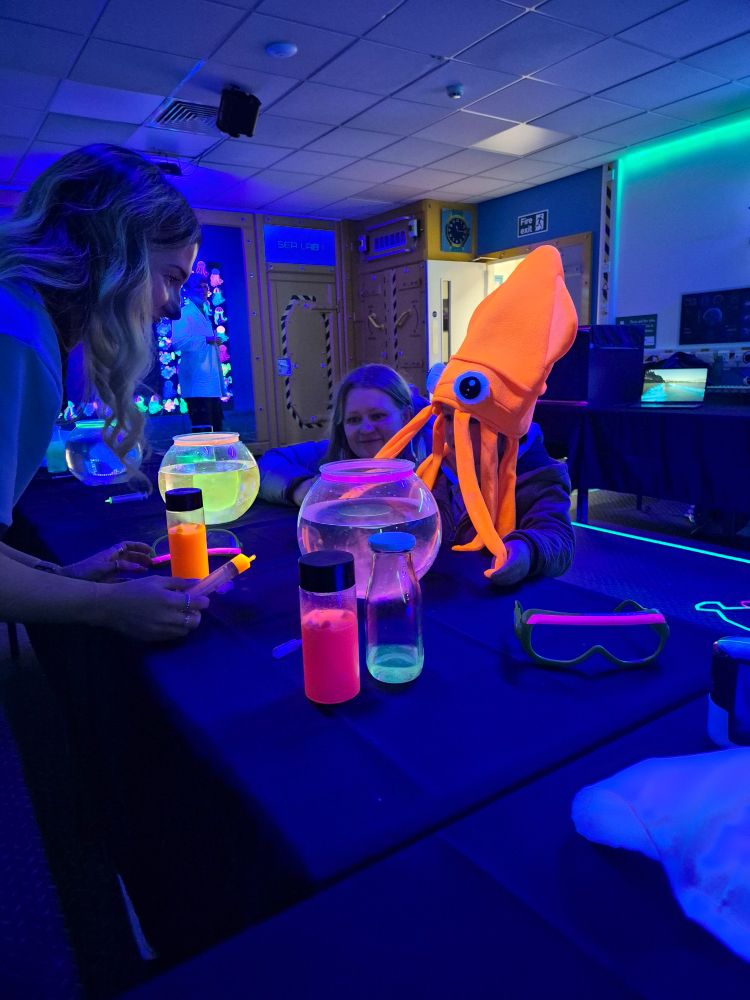 A Guide engaging with a young child wearing a bright orange jellyfish hat. The Guide is carrying out a demonstration. 