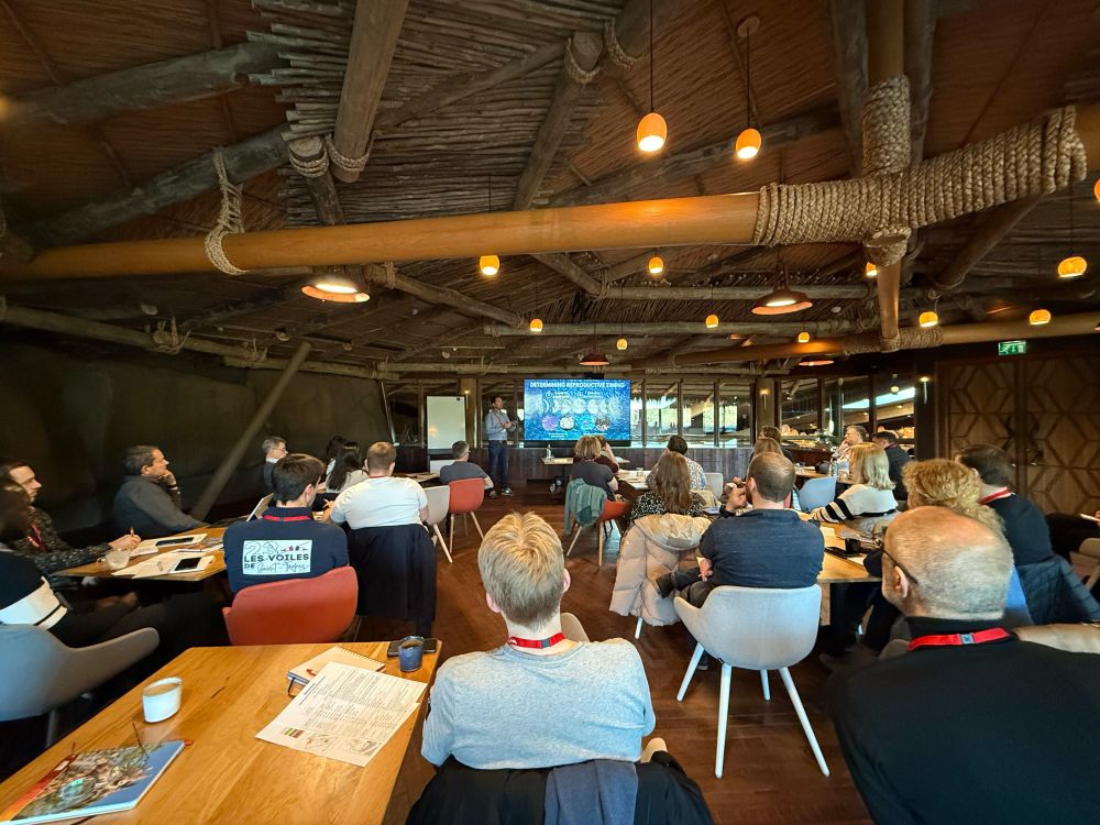 Photo taken at the back of a room filled with coral experts from around the world. They are all seated at tables and facing away from the camera towards one of the group who is stood presenting from a screen.