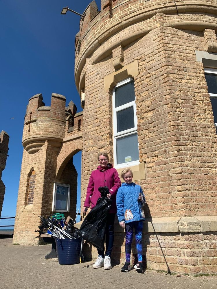 Two individuals participating in a cleanup activity stood in front of Withernsea's pier towers, holding garbage bags and litter pickers.