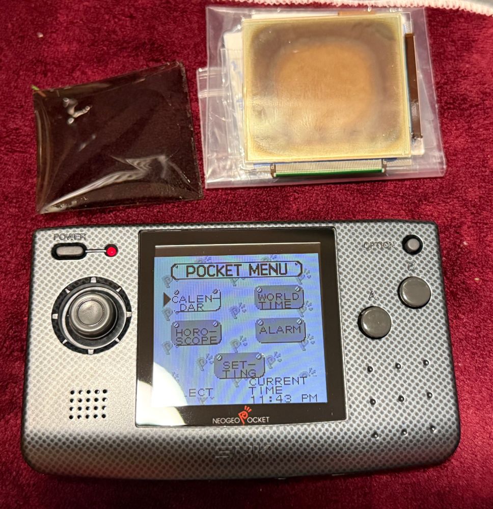 Neo Geo Pocket console with a backlit screen with the original damaged screen and polarising film above it, on a bordeaux coloured felt cloth.