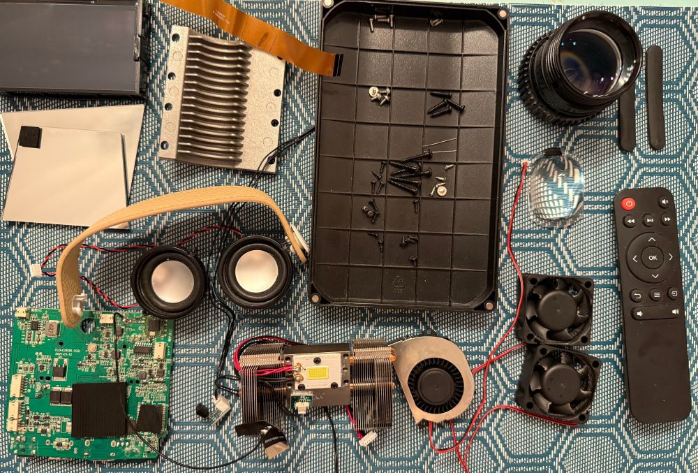 Photo of the different parts of a projector: mirrors, lenses, fans, heatsink, speakers, carry strap, screws and a remote.