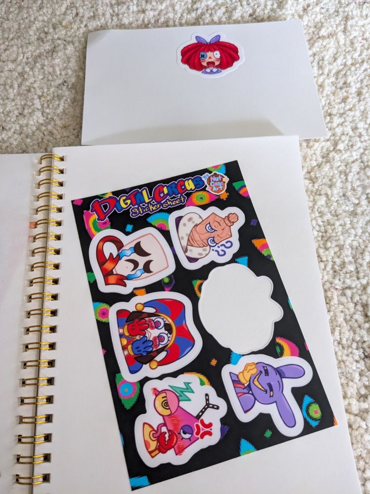 A photo of a sticker book with a sticker sheet pasted into one of the pages. It features characters from The Amazing Digital Circus, and all but one character peeled off with the sheet. Above the book is the paper the sheet was on before with the one character who didn't peel with it still on the paper: Ragatha.