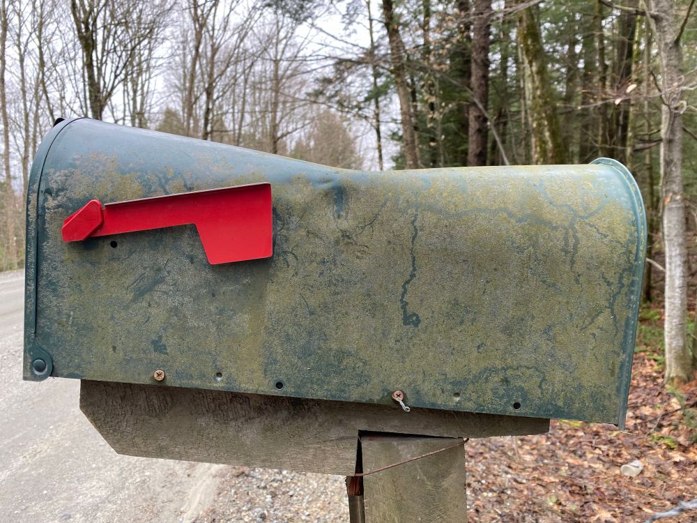 A dusty mailbox by the side of a country road, with a massive dent in the top.