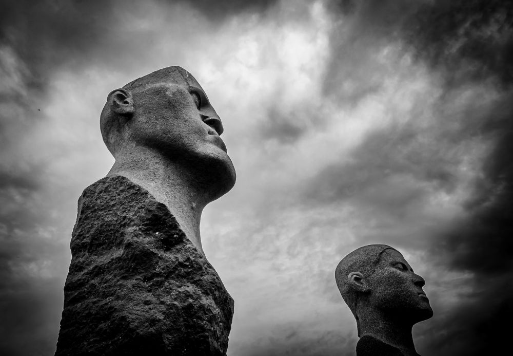 Schwarz/Weiß-Fotografie. Zwei Steinskulpturen die menschliche Köpfe darstellen, vor wolkenverhangenem Himmel.
