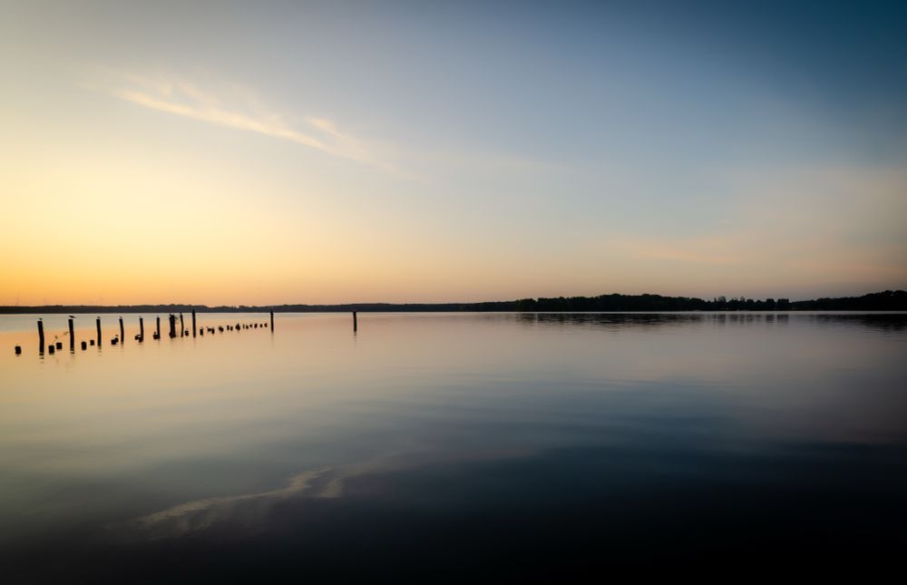 Ein blassblauer Himmel über einem fast glatten See. Das gegenüberliegende Ufer spiegelt sich leicht. Links sind Holzstämme, vielleicht Reste von einem Steg zu sehen, auf denen Möwen sitzen.
