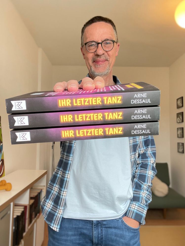 Mann mit drei Büchern. Es ist der bekannte Krimiautor Arne Dessaul, der mit der rechten Hand dreimal sein neues Werk „Ihr letzter Tanz“ in die Kamera hält, die andere Hand lässig in der Hosentasche, wie Dessaul überhaupt einen lockeren, sympathischen Eindruck hinterlässt. Bestimmt schreibt er auch so. Hach …