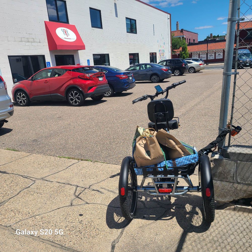 Electric trike parked in parking lot of the Aliveness Project ready for produce to be unloaded. The trike is against a chain link fence to the right cars parked to the left, the entrance is to the left with the red awning embellished with the organization's logo.