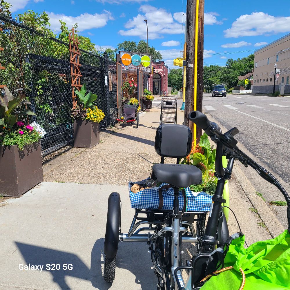 E-trike all loaded up with fresh haul of plants from Mother Earth Gardens in Longfellow neighbourhood. Trike parked on sidewalk chained to a bike parking anchor. To the left is the Mother Earth fence with planters outside all abloom  on the corner just up the sidewalk is their sign 3 bold circles, orange, teal, chartreuse, each containing one word of the store name. To the right is the asphalt street of 38th st. Just behind the store sign is the brick fascade of Lynette, the neighbourhood cafe.