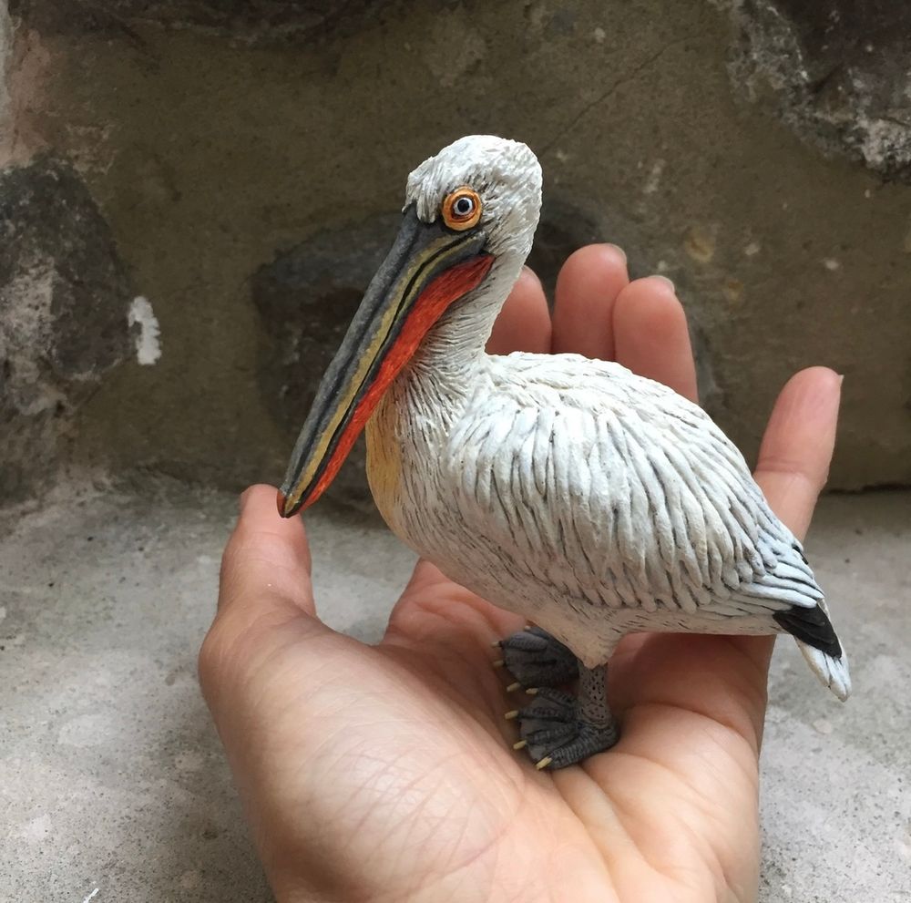 A Dalmatian pelican sculpture which is made out of air drying clay. It is painted in acrylic paint (the predominant colours are white, grey and orange) and it is stood in the palm of my hand. 