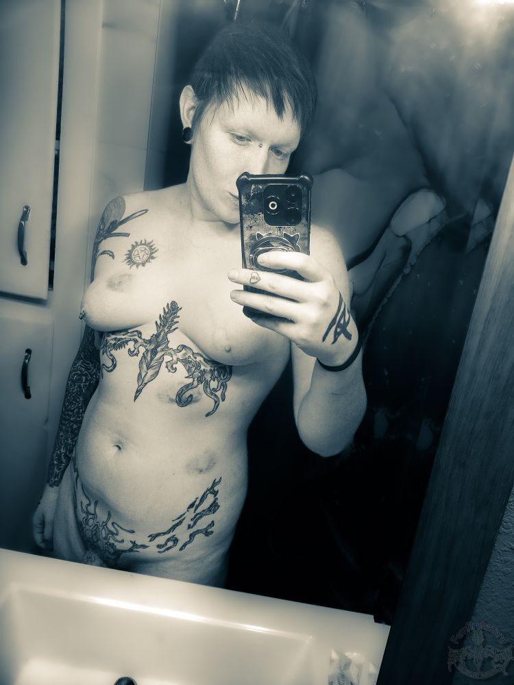 A nude tattooed transfem enby holding up a phone to take a pic in a mirror 