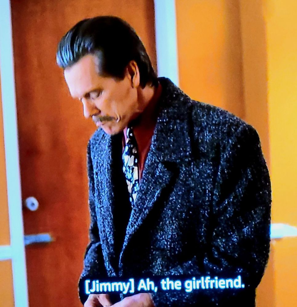 Screengrab showing a mustachioed white male in profile. The man has his next bent, wears a wooly topcoat, red shirt, and a tie-dyed kind of neck tie. White subtitle reads:

[Jimmy] Ah, the girlfriend.