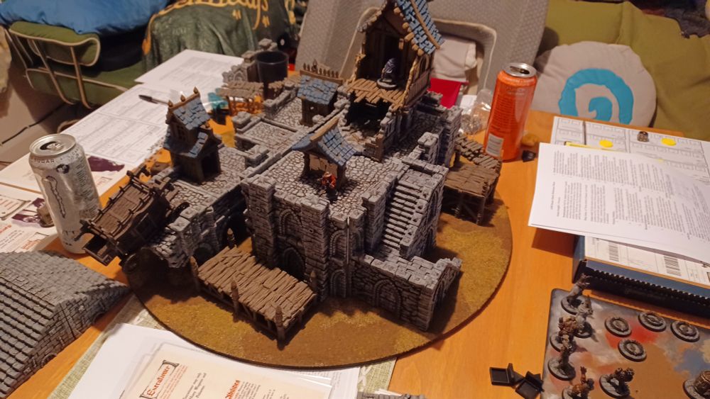 A circular game board with a large castle like structure, miniature assassins can be seen sculking around on top of it. The rest of the table is littered with rules pages and energy drinks.