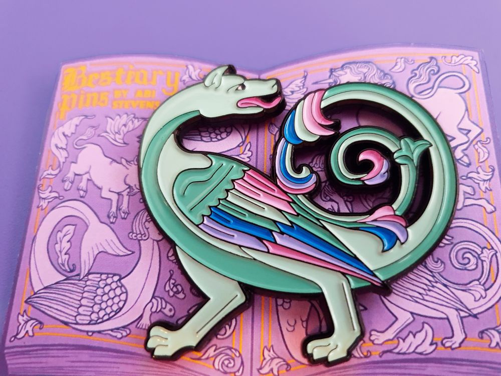 A close up photo of my dragon soft-enamel pin on a purple backing card in the shape of an open book, with monotone bestiary creatures on the pages. The pin is a black-outline mint green dragon in a bestiary inspired style, with colourful feathers and a spiral tail.