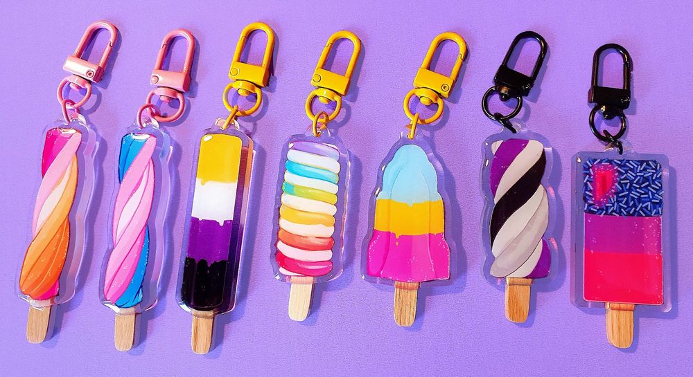 A photo of 7 different ice lolly keychain designs with different shapes and in the colours of different Pride flags: lesbian, trans, nonbinary, LGBTQIA+, pansexual, asexual, and bisexual.