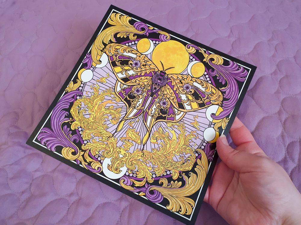 A photo of a square art print featuing my illustration of a lunar moth with leafy details in its wings and wing segements in the purple, black, yellow and white of the nonbinary pride flag, framed by a sun motif and leafy corner flourishes.