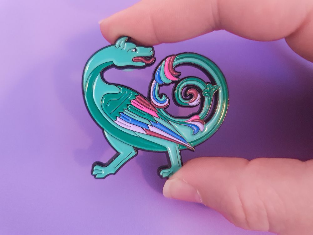 A photo of a black outlined enamel pin featuring a green dragon in the style of medieval bestiaries, with a spiral tail and feathery wing.