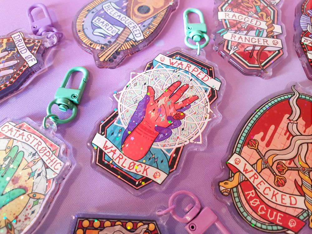 A photo of multiple acrylic keychains on a lilac background. The central design features s red hand palm up with magical sparkles appearing in the center and a larger purple transparent hand gripping it from below. There is a magic circle behind them, and a banner reading 'warped warlock'. There are other keychains around it representing other d and d classes, including catastrophic cleric, wrecked rogue, druid of disaster, and beleaguered bard. The keychains all have holographic sparkles.