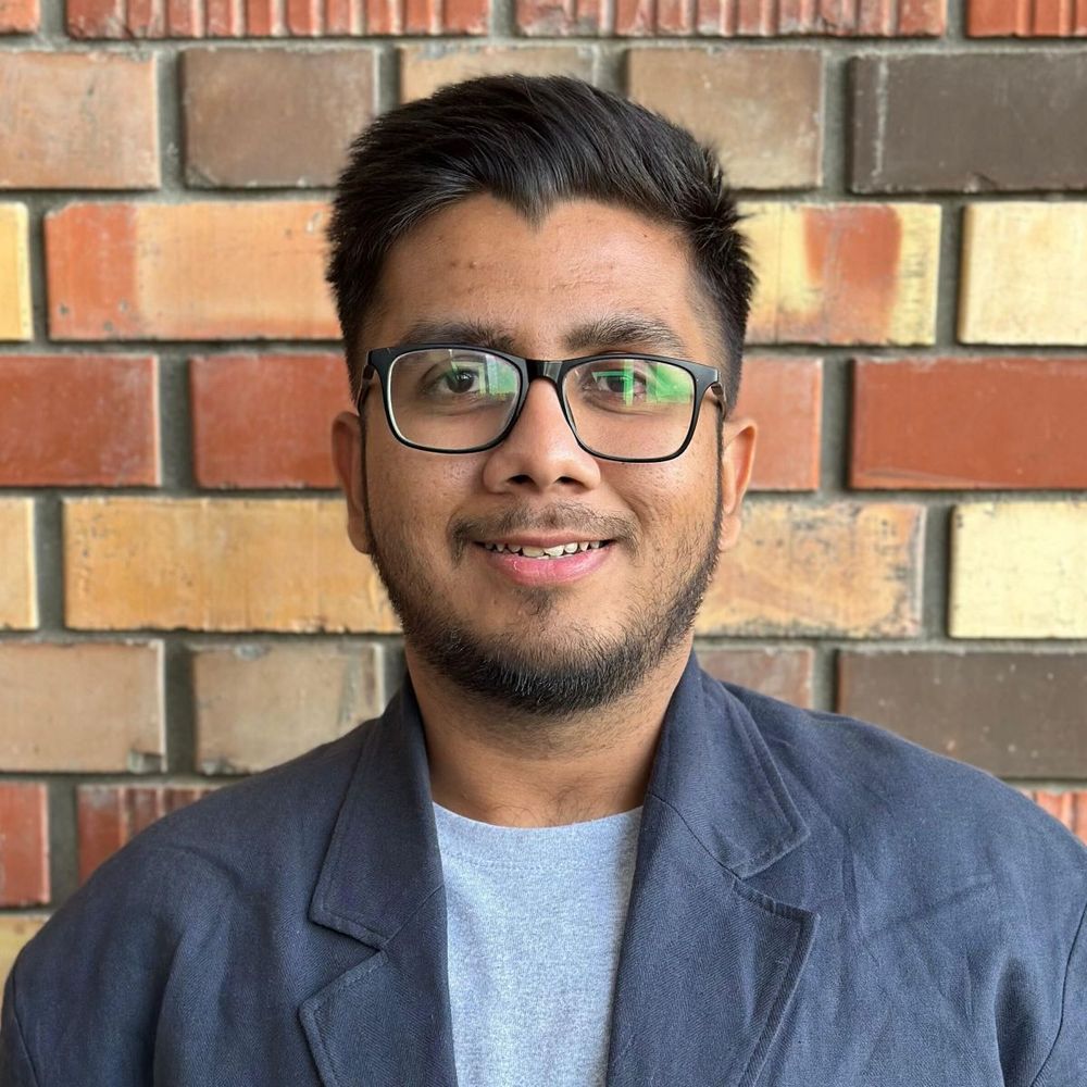 Smiling man with glasses and a beard, posing in front of a brick wall. He is wearing a gray shirt and a blue blazer.