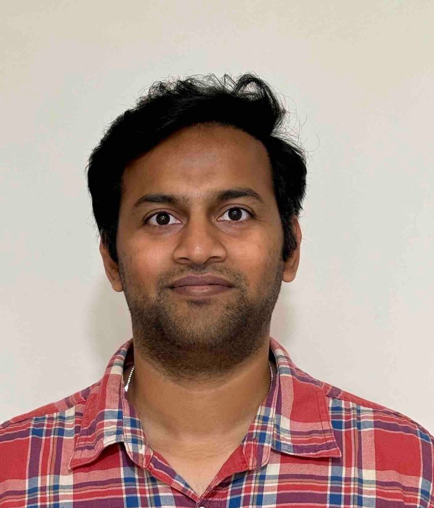 A headshot of a man with dark hair and a neatly trimmed beard, wearing a red and blue plaid shirt against a plain light background. He looks directly at the camera.