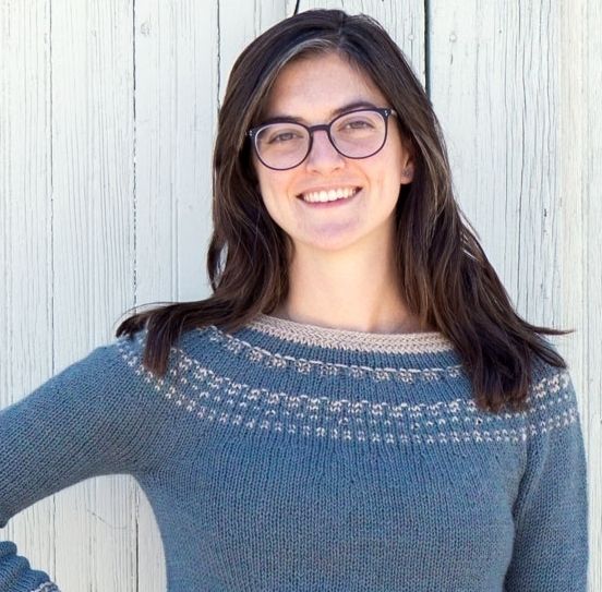 A femme person with medium length brown hair wears black-rimmed glasses and is smiling at the camera. She is wearing a blue handmade sweater with beige colorwork, a design by Tif Neilan called Sprout.