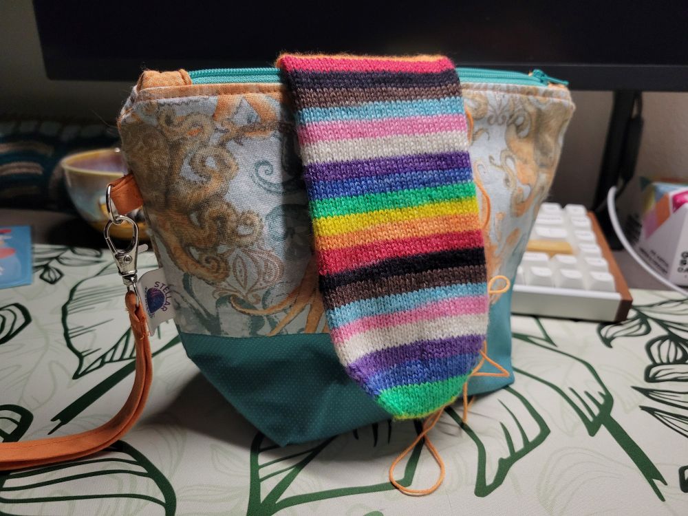A handknit sock rests on a handsewn bag with octopi on it on a computer desk.