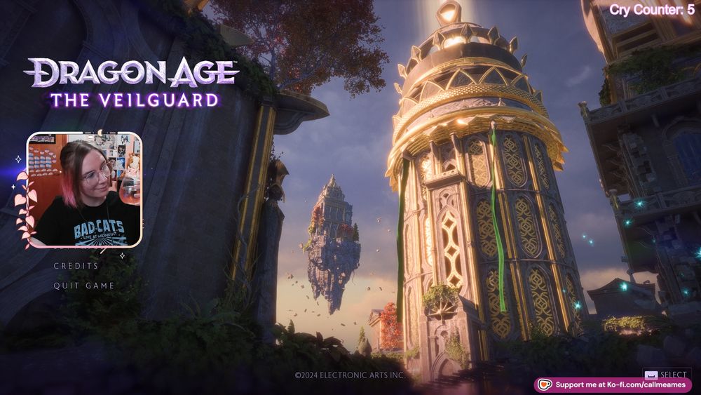 Ames' facecam over the main screen for dragon age: the veilguard Around their face cam is a light pink border with pink leaves and a crescent moon. They are smiling and holding a glass of rose wine and wearing a black shirt that says bad cats live at midnight in white font on it.