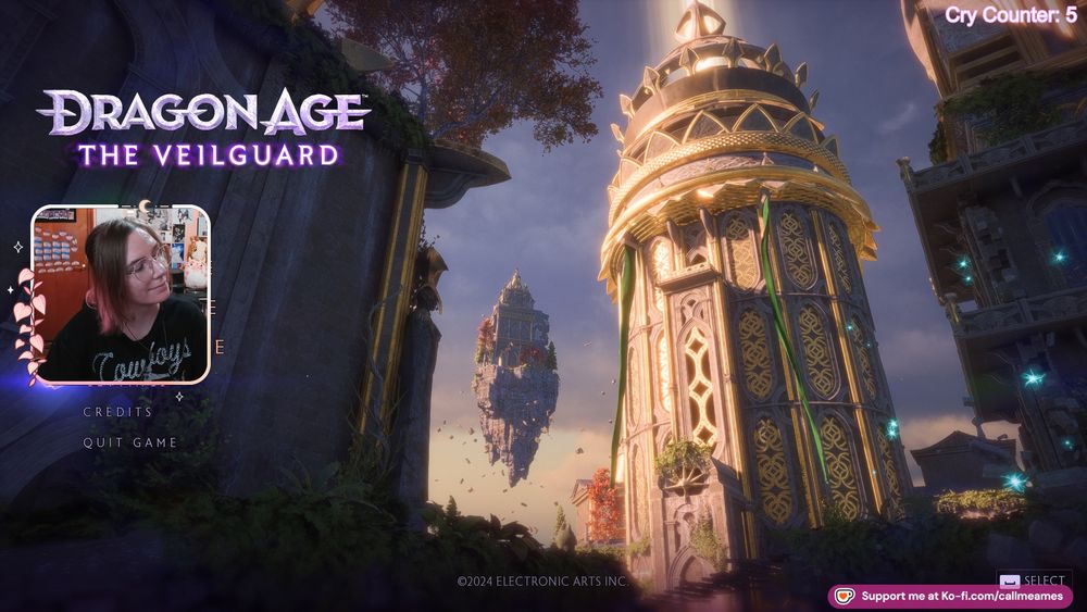 Ames' facecam over the main screen for dragon age: the veilguard. Around their face cam is a light pink border with pink leaves and a crescent moon. They are smiling with their head tilted and wearing a black shirt that says cowboys in white font.