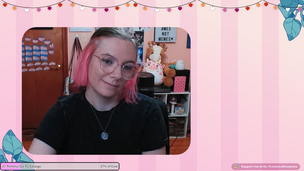 Ames' facecam over their just chatting scene. The background behind their facecam is pink striped and there are heart shaped lights at the top in the colorway of the lesbian flag. There is a kofi goal with 'Temmie go to college' on it in the lower left corner and Support me at ko-fi.com/callmeames on the right. They are smiling and wearing a black tshirt and a silver pendant necklace. Behind them is a pink wall covered in kpop posters and baldur's gate 3 art. There is pink cow squishmallow and a giraffe stuffed animal sitting on top of a cube organizer with various knicknacks.