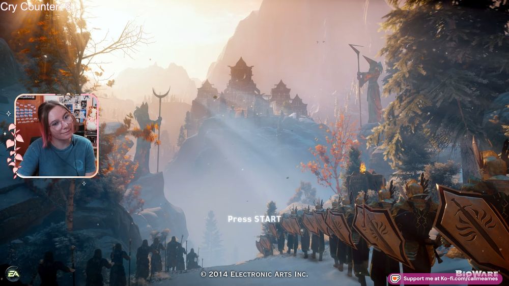 Ames' facecam over the main screen for Dragon Age: Inquisition. Around their face cam is a light pink border with pink leaves and a crescent moon. They are smiling and wearing a blue shirt.
